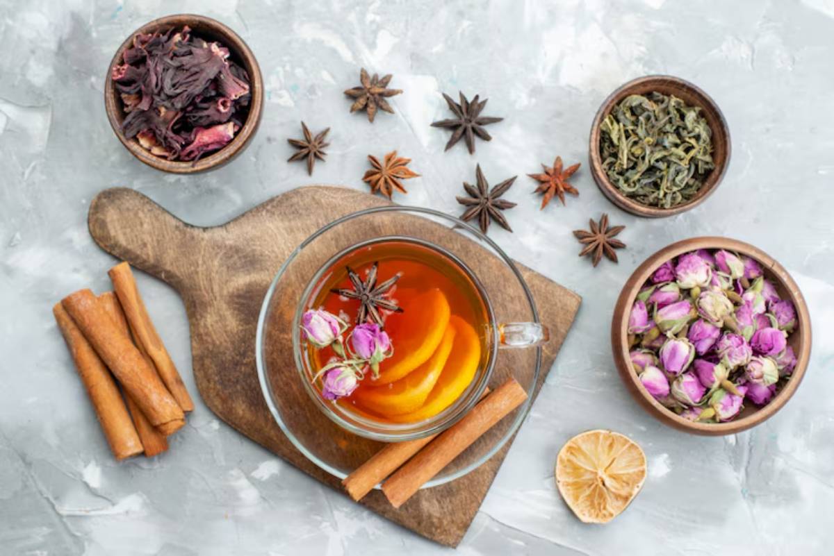 A glass of aromatic tea with orange slices and rose petals, surrounded by herbs, spices, and dried flowers on a wooden board.