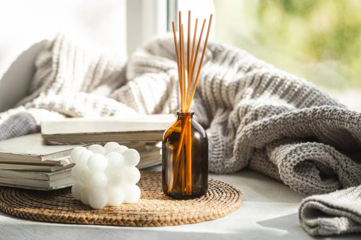 A cozy setup featuring a brown bottle with reed sticks, a white bubble candle, stacked books, and a gray knitted blanket on a woven mat.