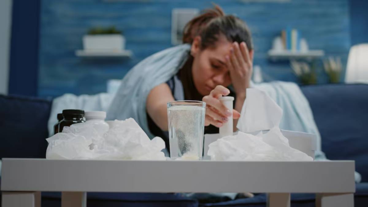 A person wrapped in a blanket sits on a couch, reaching for a tissue amid crumpled tissues and medication bottles on a table.