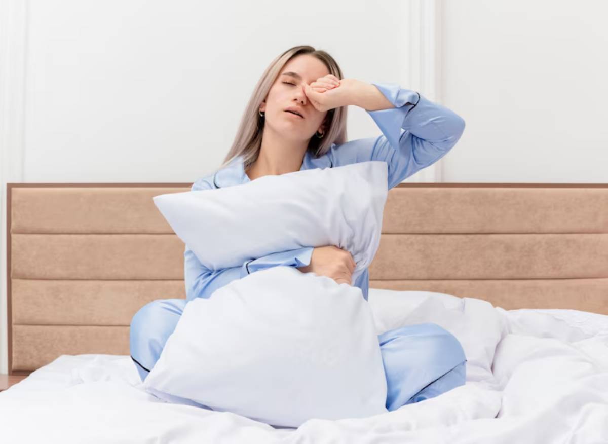 A person in light blue pajamas sits on a bed, hugging white pillows amidst rumpled white bedding.