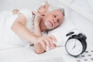 A person lies in bed, reaching for an alarm clock on a bedside table, indicating a struggle to wake up.