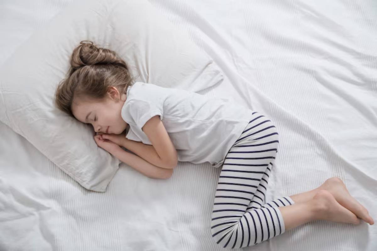 A young child curled up on a bed, resting on a white pillow, dressed in a light shirt and striped leggings.