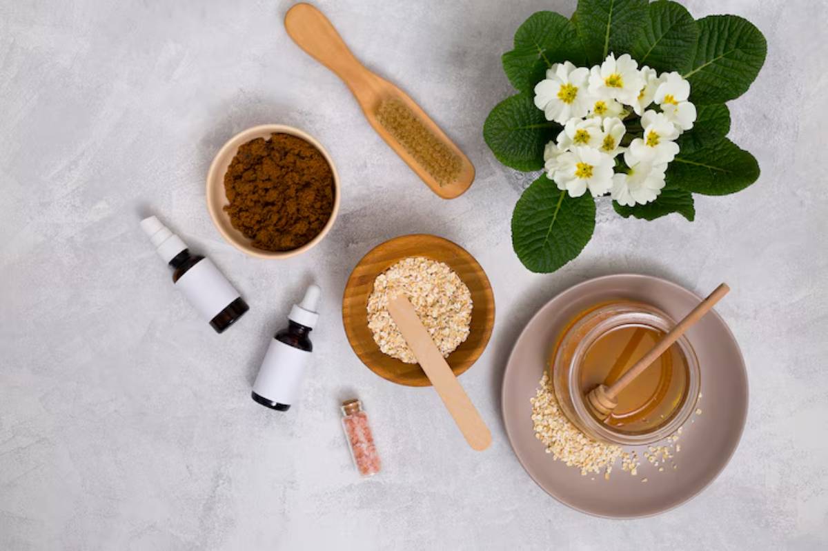 Top view of natural skincare ingredients: honey, oats, brown sugar, droppers, and a blooming white flower plant. 