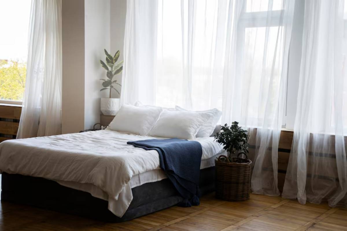 Bright and airy bedroom featuring a neatly made bed with white linens, a blue throw, plants, and sheer curtains filtering sunlight.