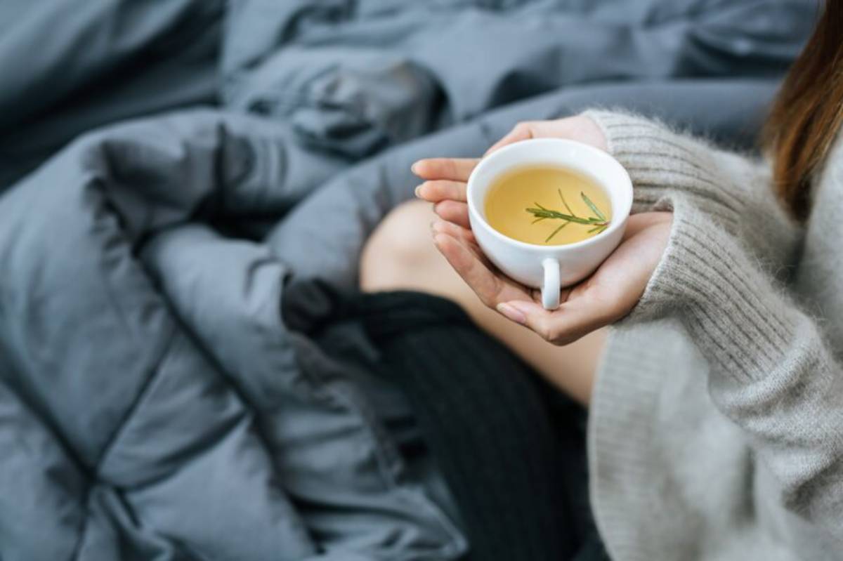 A person holds a white cup of herbal tea with a sprig, sitting on a bed covered with gray blankets, wearing a cozy sweater.