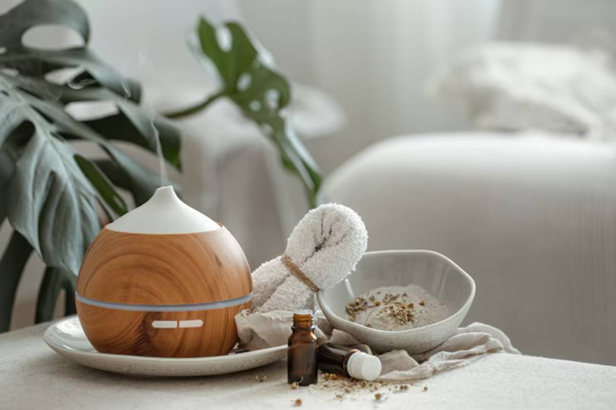 A wooden essential oil diffuser sits with a rolled towel, bowl of herbs, and oil bottles, creating a calming wellness scene.