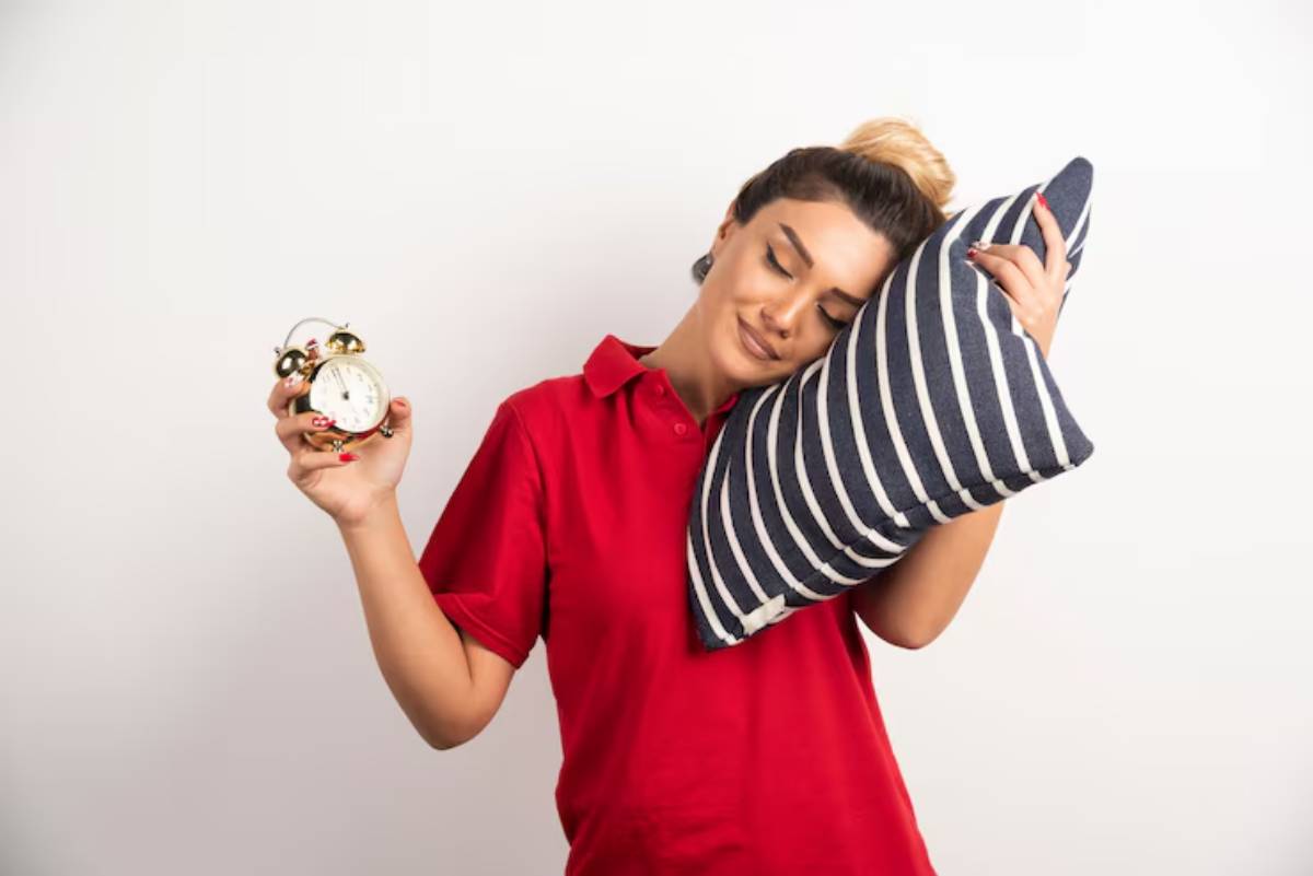 A person in a red shirt holds an alarm clock in one hand and a striped pillow in the other against a plain white backdrop.