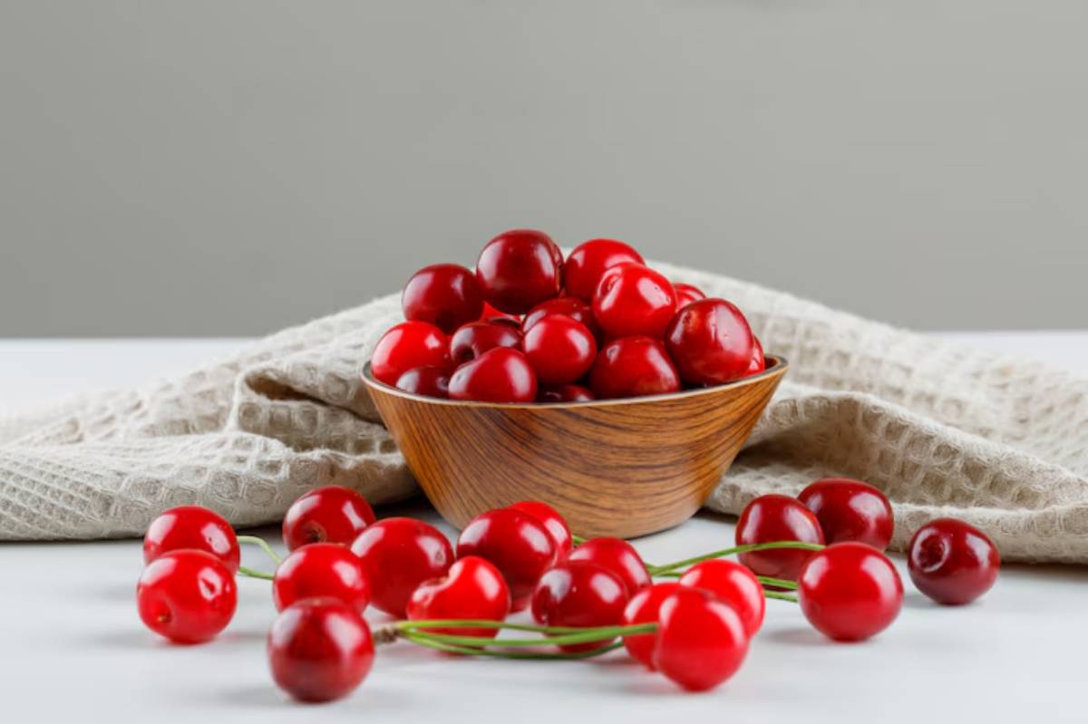 A wooden bowl overflowing with shiny red cherries, surrounded by more cherries on a white surface, draped with a textured beige cloth.