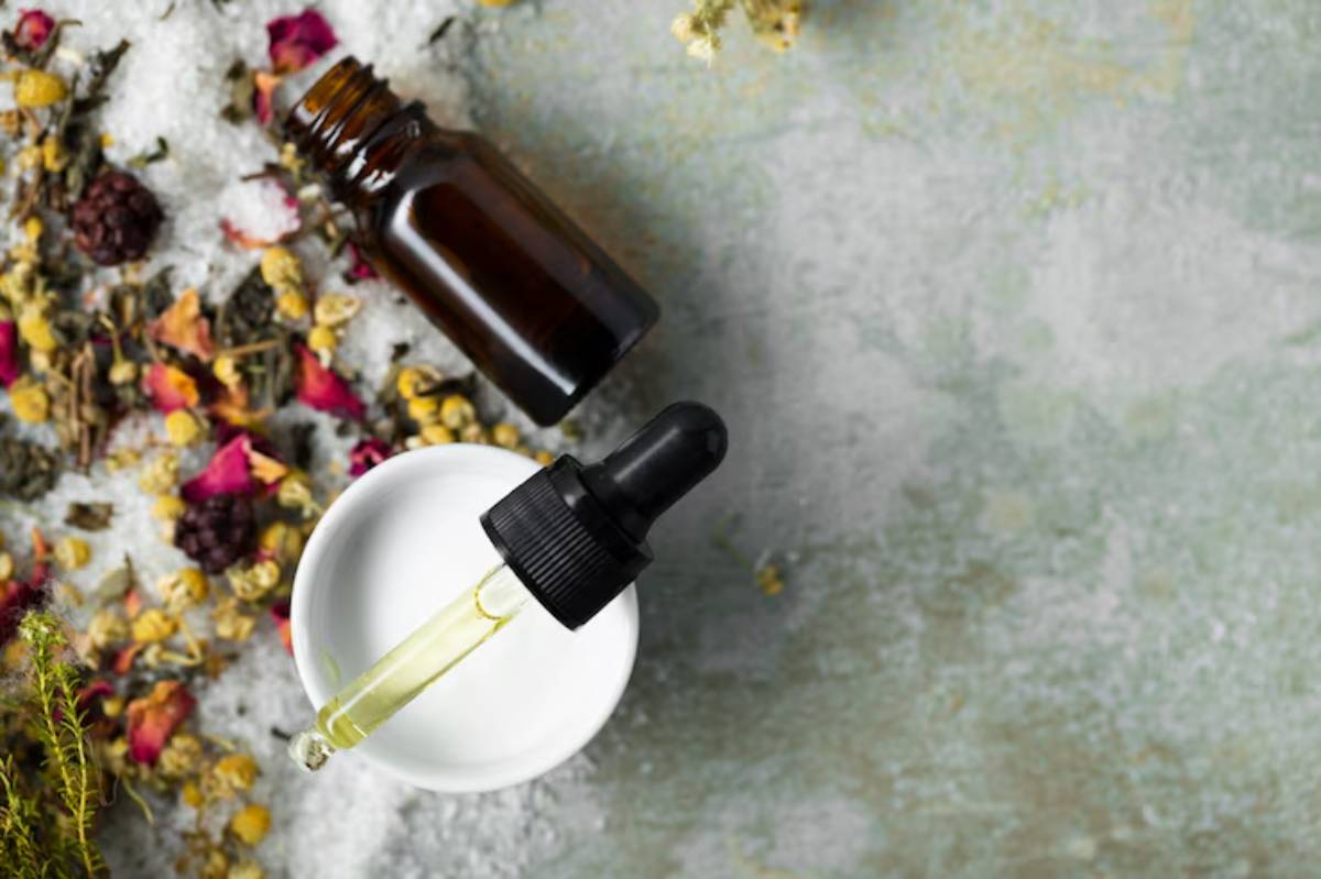 A dropper filled with yellow oil hovers above a white bowl, surrounded by herbs, flowers, and textured salt on a muted background.
