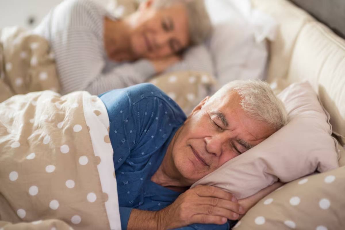 An elderly person lying in bed under a polka-dotted blanket, resting peacefully beside another person.