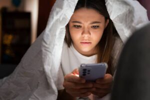 A person wrapped in a white blanket is intently looking at their smartphone in a dimly lit room.