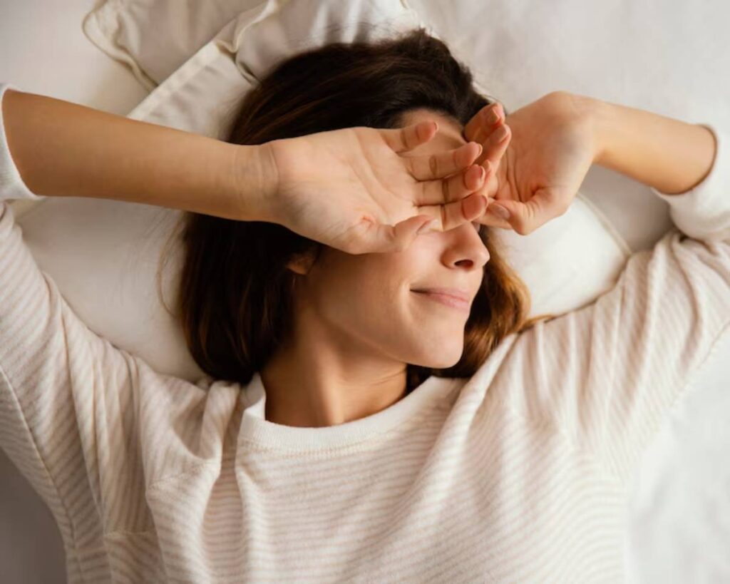 A person lies on a bed, resting their arms above their head, wearing a comfortable, light-colored striped sweater. Soft bedding surrounds them.