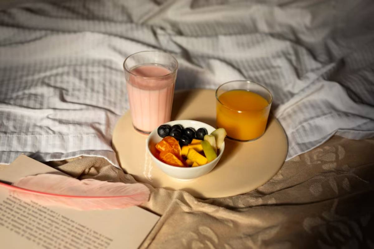 A breakfast tray on a bed featuring a bowl of mixed fruits, a glass of orange juice, and a pink smoothie, with a book and feather nearby.