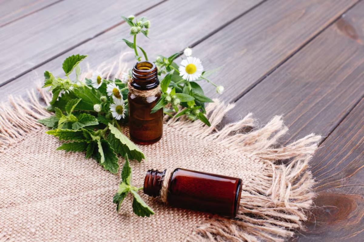 Two amber essential oil bottles surrounded by fresh herbs and white flowers on a burlap mat over a wooden surface.
