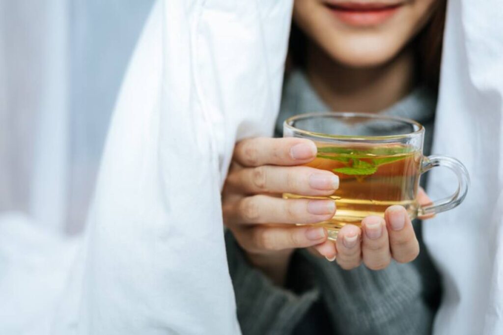 A woman holds a glass cup of herbal tea with mint, cozy under a white blanket, smiling gently.