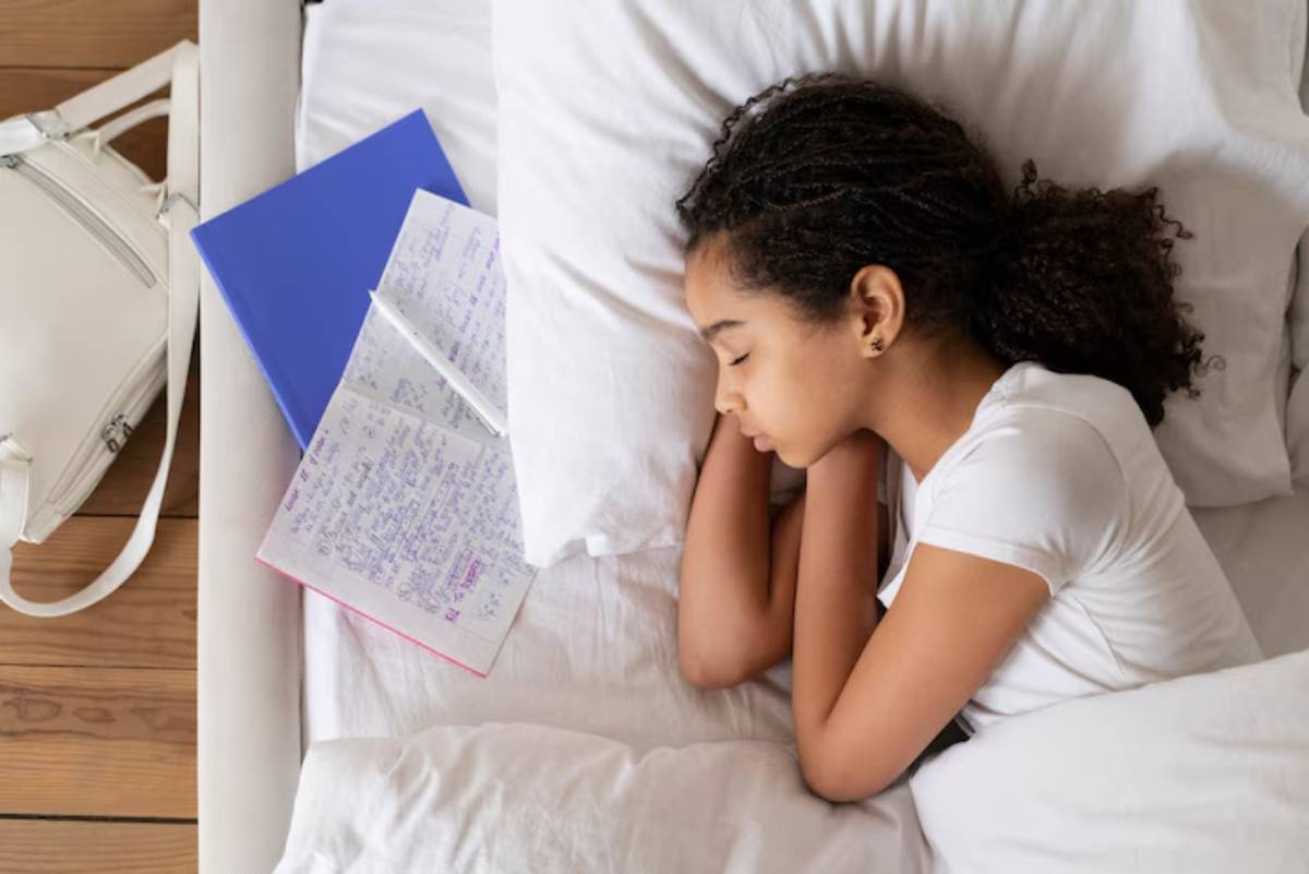 A young woman with curly hair lies on her side in bed, surrounded by notebooks and a white bag. The room has a cozy, serene atmosphere.