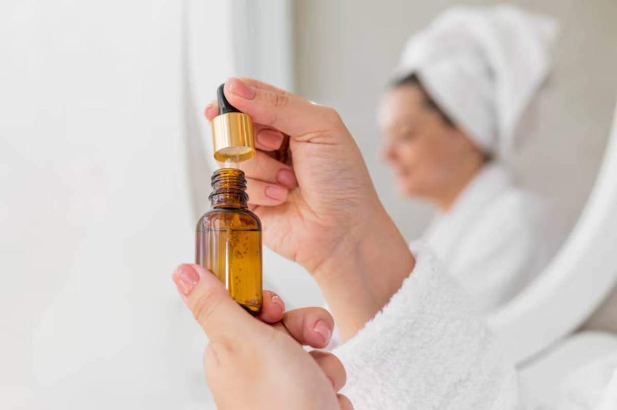 A person holds a dropper bottle of amber essential oil, preparing to apply it, with a soft-focus background of a bathroom setting.