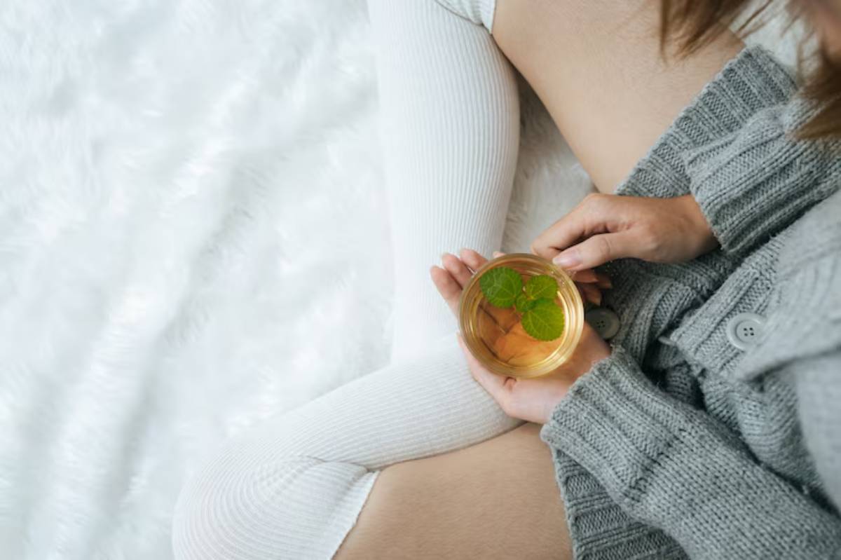 A person in a cozy gray sweater holds a glass of herbal tea with mint, sitting on a soft, white textured surface.