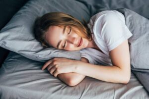 A person lies curled up in bed on gray sheets, resting their head on a pillow, with arms gently crossed around their body.