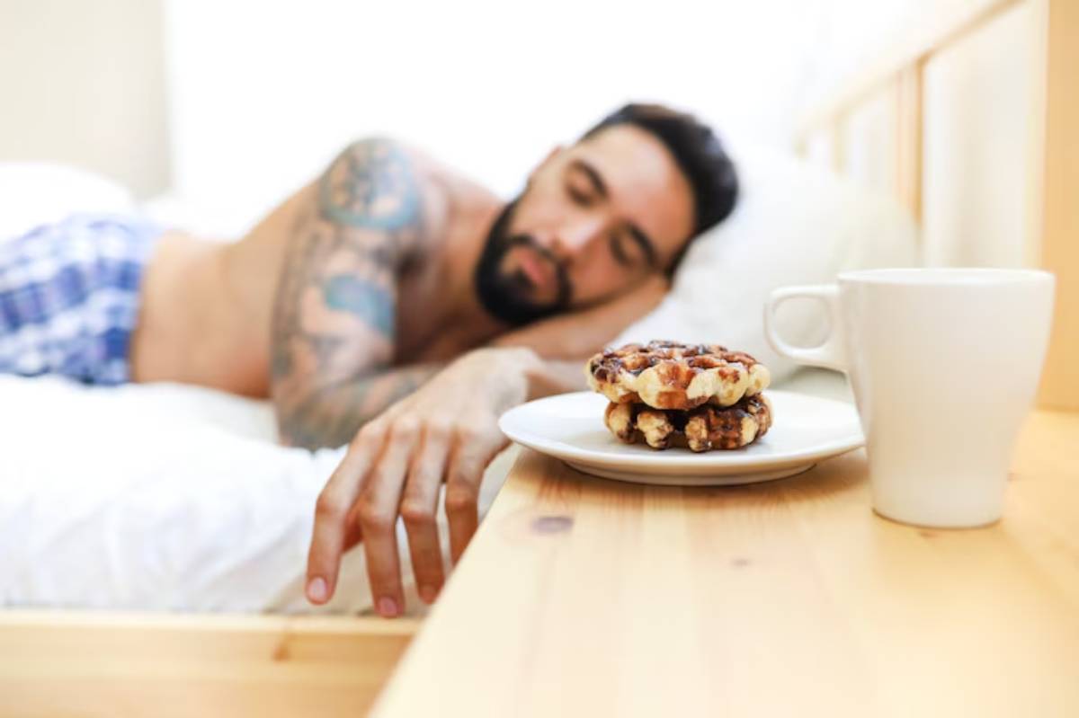 A hand reaches for a plate with two cookies and a white coffee mug on a wooden bedside table, with a bed in the background.