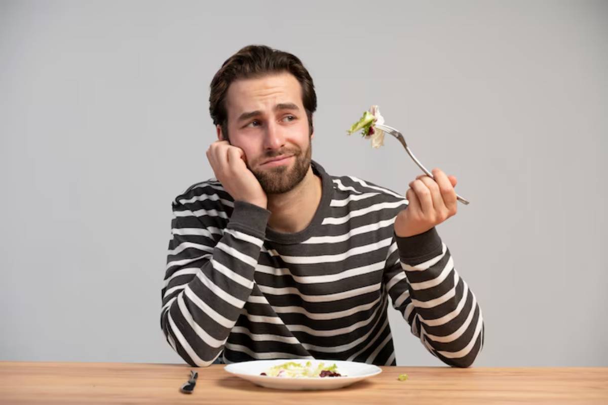 A person in a striped shirt rests their chin on one hand while holding a fork with salad, sitting at a wooden table against a gray backdrop.