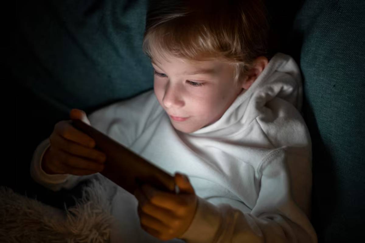 A child in a white hoodie sits on a couch, focused on a smartphone screen illuminated in a dimly lit room.