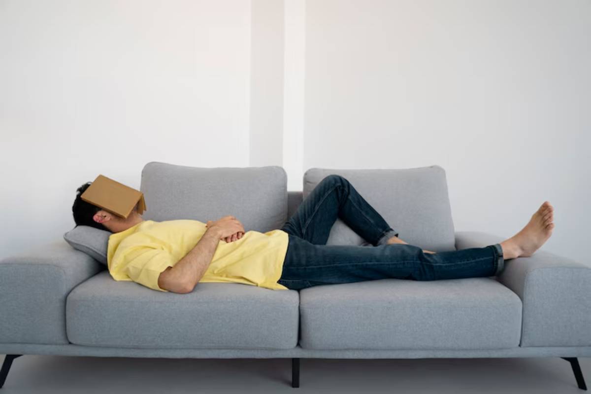A person in a yellow shirt lies on a gray couch with a cardboard box over their face, appearing relaxed and casual.