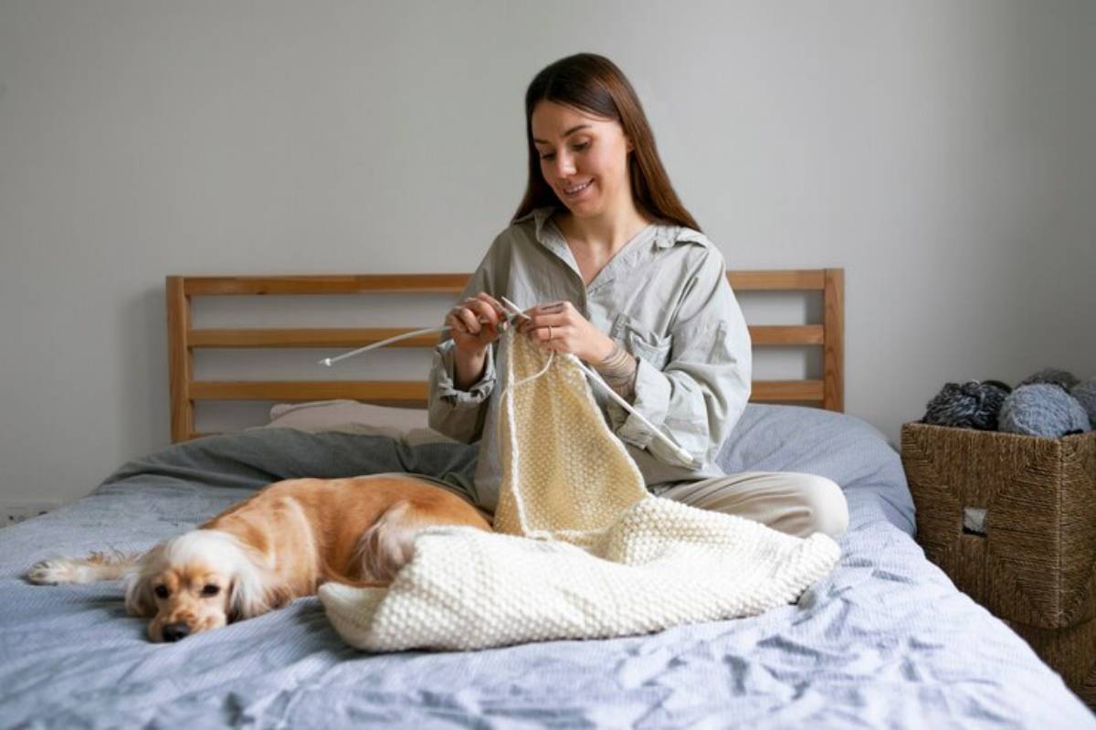 A person in a light shirt is knitting on a bed, with a golden retriever relaxing beside them and a basket of yarn nearby.