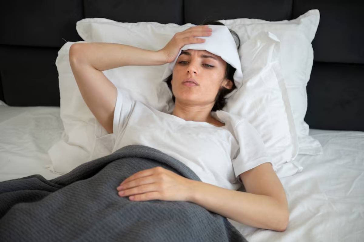 A person lying in bed, covered with a gray blanket, holding their forehead, appearing unwell and resting on white pillows.