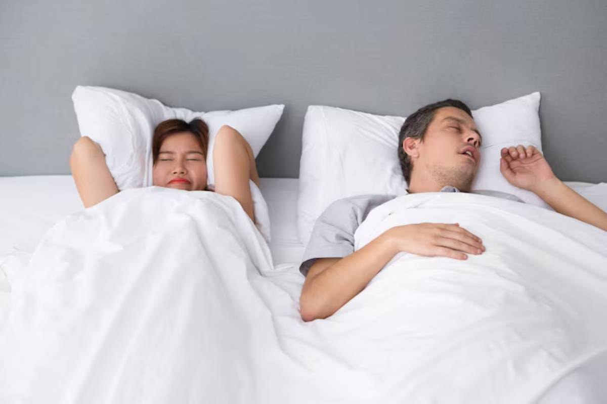 A couple lies in bed under white blankets, facing away from each other, with crisp white pillows against a grey wall.