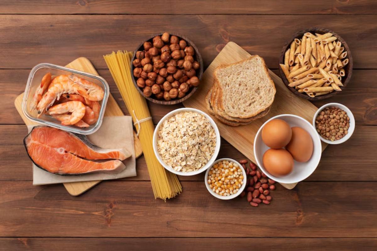An assortment of healthy foods: shrimp, salmon, pasta, eggs, whole grain bread, oats, nuts, and legumes on a wooden surface.