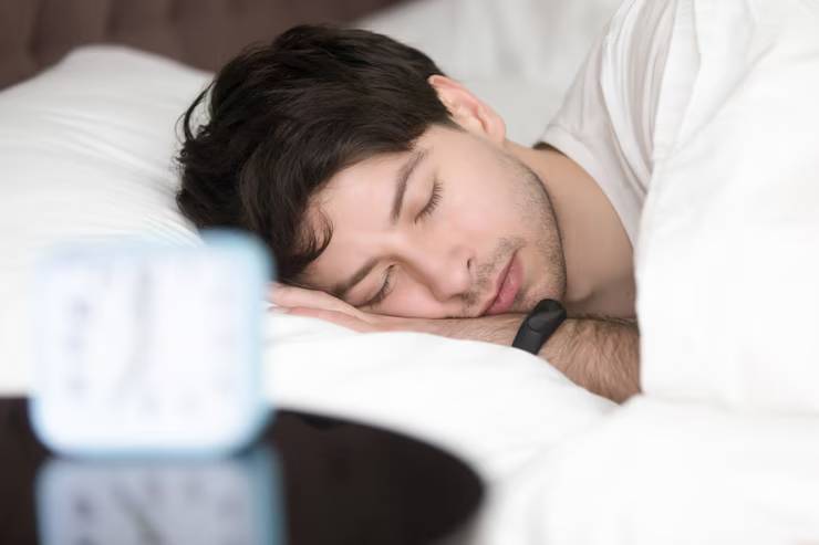 A person rests comfortably in bed, surrounded by fluffy pillows, with a bedside clock displaying the time nearby.