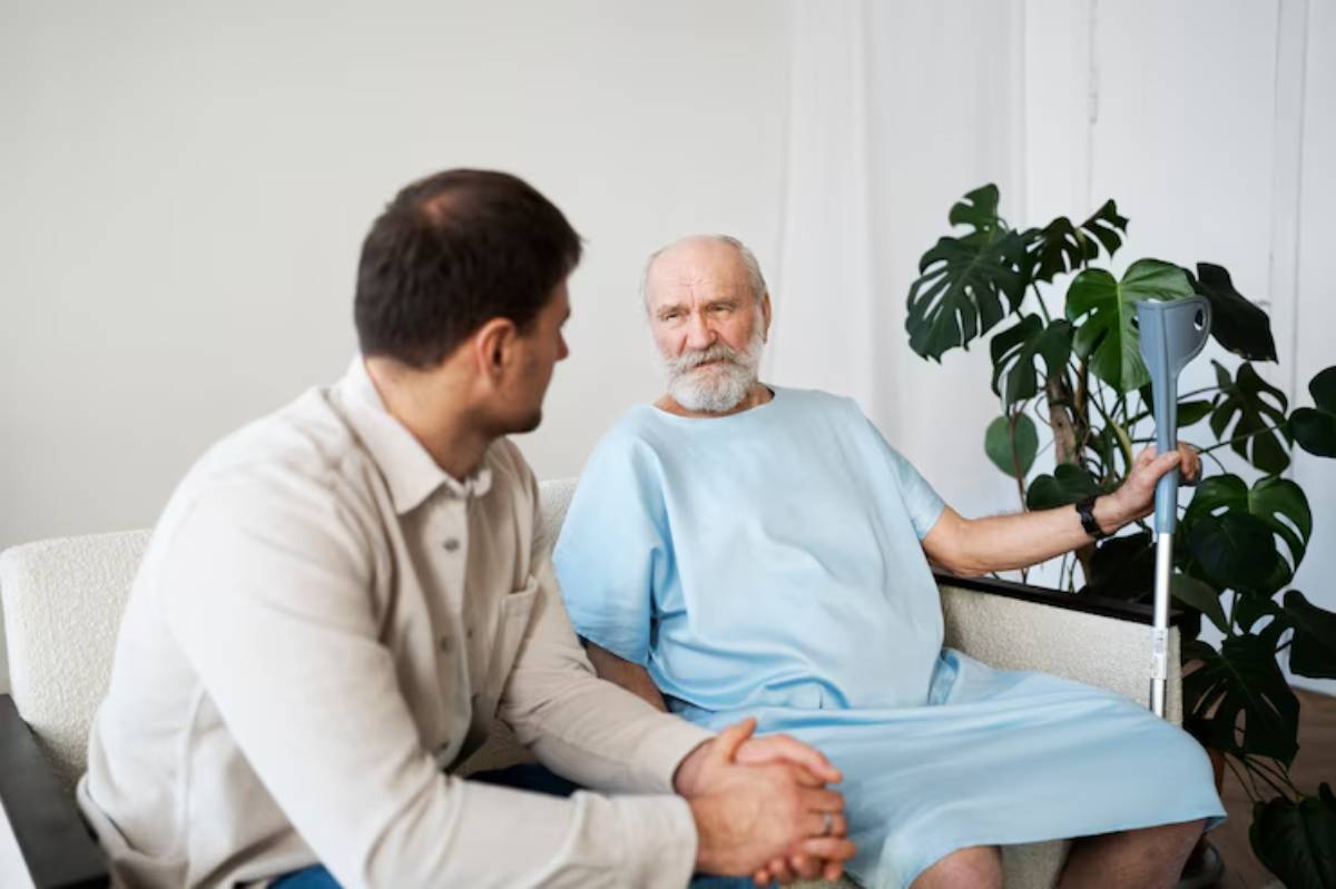 A man in a light shirt converses with a seated elderly person wearing a blue hospital gown, surrounded by indoor plants.