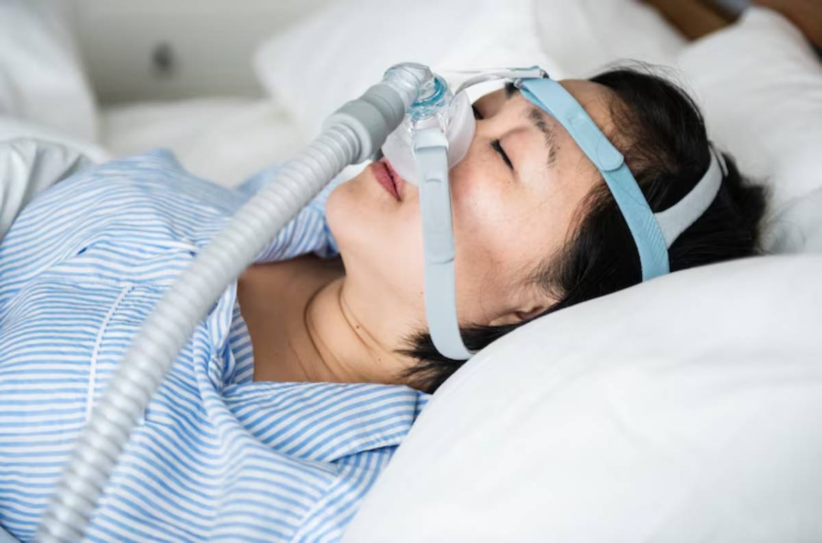 A person wearing a breathing device lies on a bed surrounded by white pillows, dressed in a blue and white striped shirt.