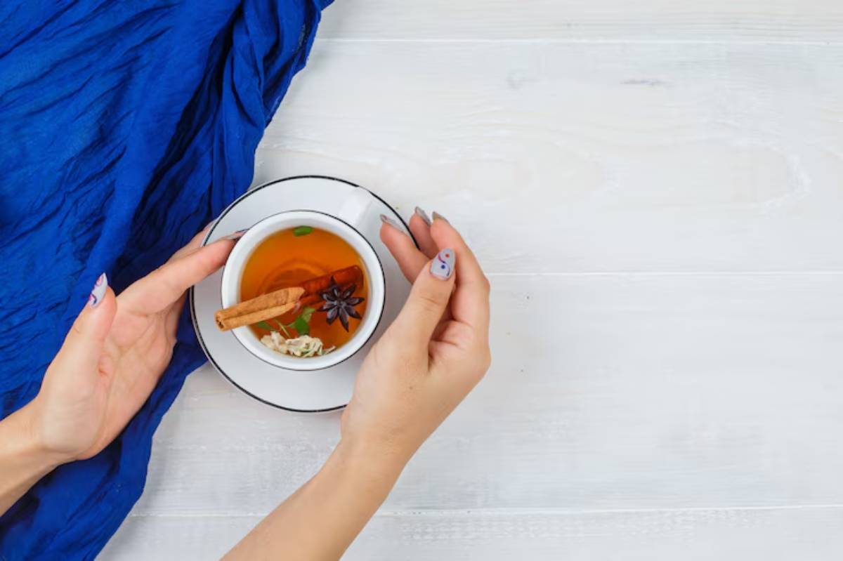 A pair of hands gently cradles a cup of spiced herbal tea, adorned with cinnamon and star anise, beside a vibrant blue fabric.