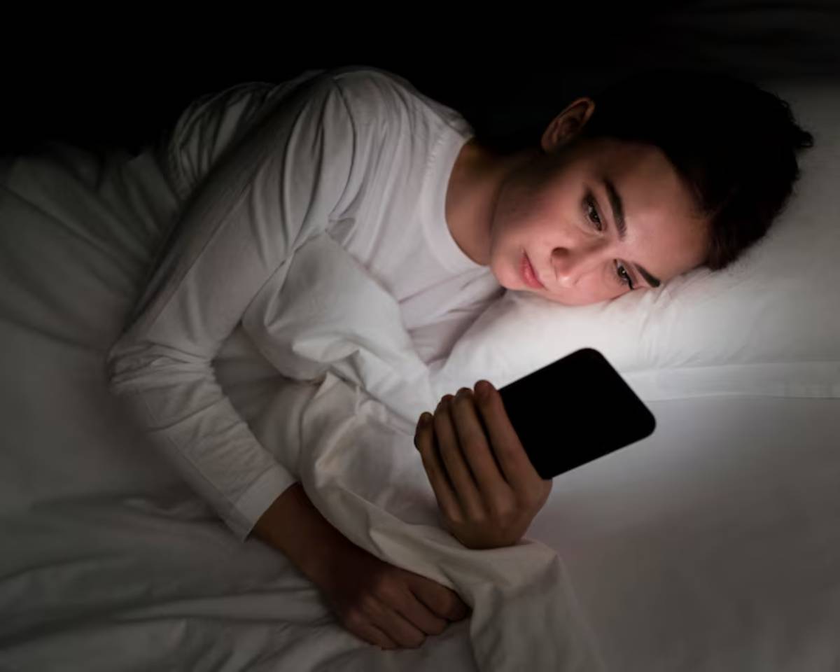 A person lies in bed under a white blanket, holding a smartphone that casts a soft glow in a darkened room.